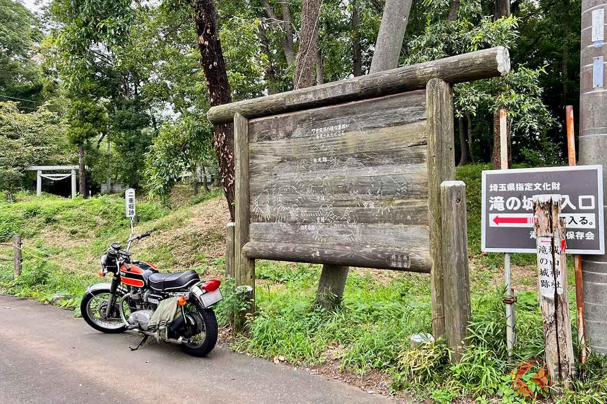 バイクで住宅地のすぐ脇を抜け、「滝の城跡」入口に到着。木製の案内板には本丸跡や空堀などの配置に加え、城よりはるか以前の横穴墓群が発見された場所も示されている。バイクを駐車場へ移動して散策開始